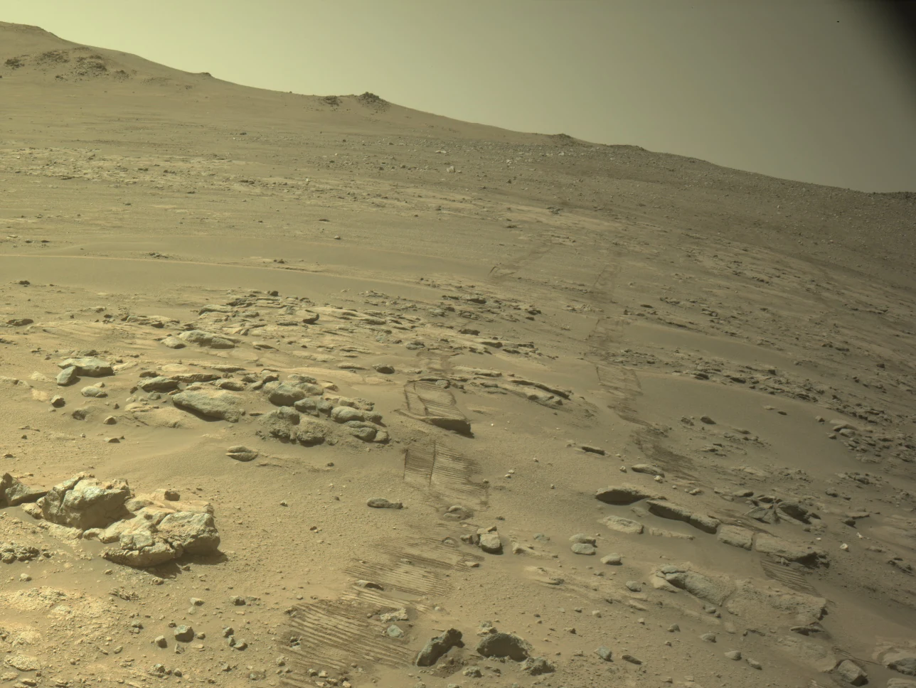 Traces de roues de Perseverance sur un sol rocheux de Mars, avec des reliefs à l’horizon.