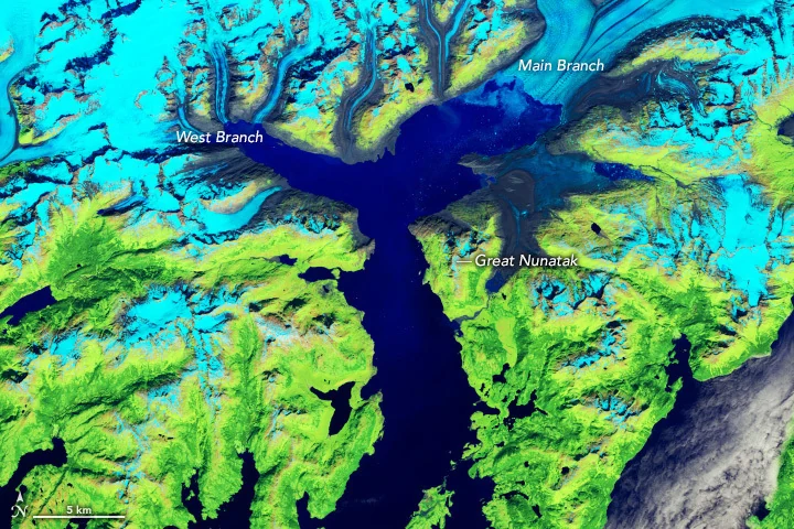 Un glacier cyan débouche dans une baie sombre, entouré de reliefs bruns.